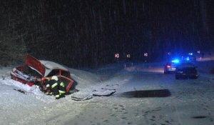 Widoczna zaśnieżona droga. Po lewej stronie na poboczu czerwony samochód uszkodzony z przodu. W tle widoczne światła błyskowe radiowozu i straży pożarnej.