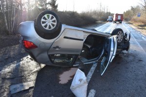 Widoczny leżący na dachu na jezdni samochód koloru srebrnego.