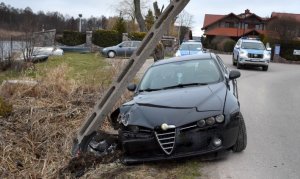 Na zdjęciu widoczny samochód marki Alfa Romeo stojący przodem na drodze. Po lewej przewrócił się na ten pojazd betonowy słup energetyczny.