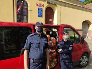 Na tle czerwonego samochodu dostawczego z otwartymi drzwiami po prawej stoi policjant, a po lewej policjantka. Oboje są umundurowani.