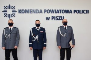 Trzech umundurowanych policjantów stojących na tle ściany z napisem Komenda Powiatowa Policji w Piszu.