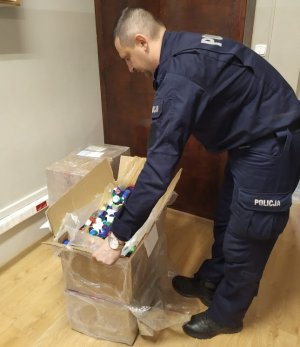 Po prawej stronie umundurowany policjant. Jest pochylony nad kartonem stojącym na podłodze. pudełko jest otwarte. Są w nim nakrętki.