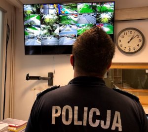 Umundurowany policjant stojący tyłem obserwuje ekran, na którym widać obrazy z miejskiego monitoringu.