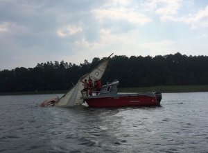 Widać jezioro i przewróconą żaglówkę, którą służby starają się podnieść z wody.