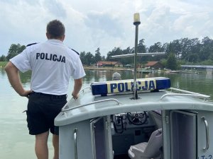 Łódź policyjna pływająca po jeziorze. Na niej stojący tyłem policjant, a w tle plaża.