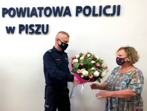 Umundurowany policjant stojący po lewej wręcza bukiet kwiatów stojącej po prawej kobiecie.