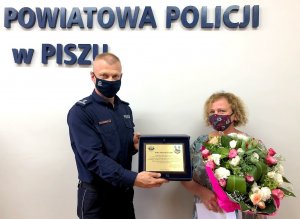 Umundurowany policjant stojący po lewej wręcza pamiątkowy ryngraf stojącej po prawej kobiecie.