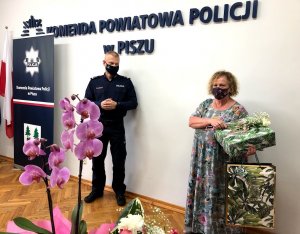 Za kwiatem widocznym na pierwszym planie dalej widać umundurowanego policjanta stojącego po lewej i kobietę stojąca po prawej.