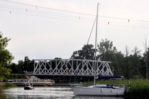Na pierwszym planie widać łódź żaglową na rzece. Po lewej policyjna łódź. W tle widoczny jest most kolejowy.