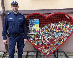 Po lewej stoi umundurowany policjant. Po prawej czerwony pojemnik w kształcie serca, w którym są nakrętki.