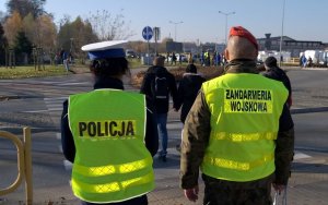 Przed przejściem dla pieszych tyłem stopi po lewej umundurowana policjantka, po prawej żołnierz Żadarmerii Wojskowej.