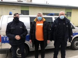 Na tle oznakowanego radiowozu stoi dwóch umundurowanych policjantów w maseczkach, a po środku mężczyzna także w maseczce.