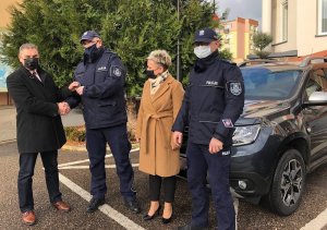 Na parkingu stoi nieoznakowany radiowóz marki Dacia koloru granatowego. Przed nim cztery osoby w tym dwóch policjantów w  mundurze.