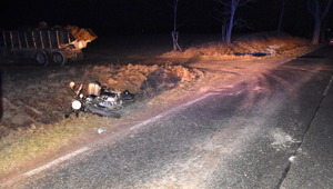 Jest ciemno. Skrzyżowanie dróg. na pierwszym planie widoczny przewrócony motorower. W tle po lewej stronie w polu stoi ciągnik rolniczy z naczepą.