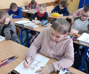 Uczniowie siedzą w szkolnych ławkach i rysują na kartkach Polę.