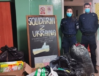 W pomieszczeniu po prawej stoi umundurowany policjant i policjantka.  Po lewej tablica z napisem &quot;Solidarni z Ukrainą&#039; Przed nimi popakowane worki i kartony.