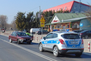 Na pierwszym planie widoczny oznakowany radiowóz stojący na drodze. Ustawiony jest tyłem. na drugim planie widać stojący na drodze samochód osobowy w kolorze bordowym.