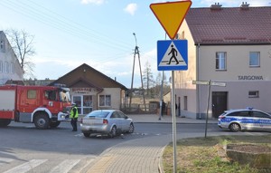 Skrzyżowanie ulic. Po lewej na drodze stoi samochód straży pożarnej. Po prawej oznakowany radiowóz. Miedzy nimi widać umundurowanego policjanta, a na pierwszym planie samochód koloru srebrnego ustawiony tyłem