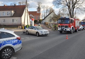 Na pierwszym planie widoczny fragment oznakowanego radiowozu. Dalej po lewej samochód osobowy koloru srebrnego stojący na skrzyżowaniu. W tle widać samochód strażaków.