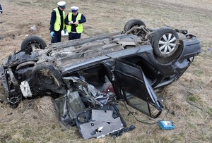 Na łące leży przewrócony samochód. Przy nim dwaj umundurowani policjanci.