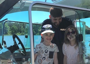 Policyjna łódź. W środku umundurowany policjant i dwójka dzieci pozujących do zdjęcia, w tym dziewczynka po prawej ma twarz wymalowana na kotka.