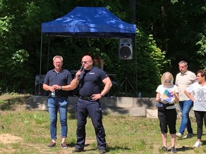 Po prawej grupka osób. Po środku umundurowany policjant trzymający w ręku mikrofon. Po lewej stronie stoi drugi mężczyzna w ubiorze cywilnym.