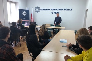Sala odpraw w KPP Pisz. na krzesłach siedzi młodzież. Przed nimi stoi umundurowany policjant- Komendant, który przemawia do młodzieży.