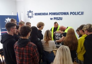 Sala odpraw KPP Pisz. Dwóch policjantów w kamizelkach odblaskowych prezentuje młodzieży walizkę technika kryminalistycznego.