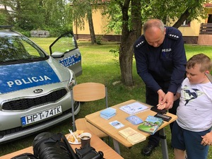 Po lewej oznakowany radiowóz. Po prawej policjant robi chłopcu odciski palców.