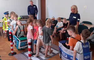 Dzieci bawią się w sali w miasteczku ruchu drogowego pod okiem policjantki stojącej po prawej,