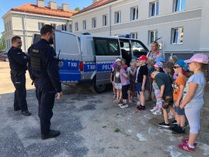 Po prawej stoją dzieci, po środku widoczny radiowóz z otwartymi drzwiami, a po lewej stoi dwóch policjantów.