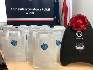 Na stole stoją niewielkie białe torebki z logo akcji. Po prawej deska do pływania dla dzieci z napisem policja i czerwona dużą kokardą. W tle baner Komendy Powiatowej Policji w Piszu.