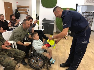 Po prawej umundurowany policjant, który wręcza odblaskowy worek chłopcu siedzącego po lewej na wózku inwalidzkim.