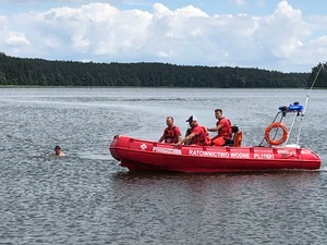 Na jeziorze po prawej widoczna pomarańczowa łódź, którą płyną ratownicy. Po lewej w jeziorze widać tonącego człowieka.