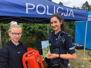 Na tle namiotu z napisem POLICJA, po prawej stoi umundurowana policjantka, która wręcza dziewczynie stojącej po lewej ulotkę o bezpiecznych wakacjach i bojkę ratunkową.