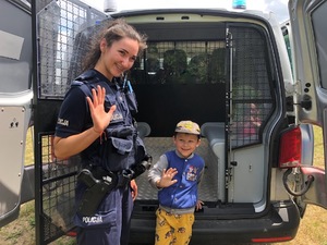 Z lewej umundurowana policjantka, a po prawej chłopiec. Oboje stoją na tle radiowozu i uśmiechają się.