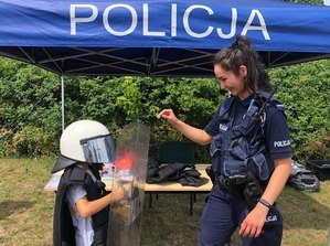 Po prawej policjantka, która ubiera policyjny kask dziecku  widocznemu po lewej. Oboje stoją na tle namiotu z napisem POLICJA.