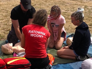 Na plaży tyłem odwrócona siedzi kobieta- ratownik wodny. Przed nią mężczyzna, który pochyla się nad fantomem, a przy nim dwie dziewczynki.