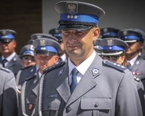 Na zdjęciu wizerunek policjanta w mundurze wyjściowym stoją na dworzu na tle innych policjantów.