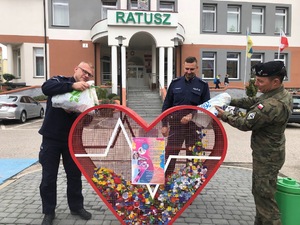 Pojemnik w kształcie serca przed Urzędem Miejskim w Orzyszu. Po lewej stoi policjant, a po prawej żołnierz. Panowie wsypują nakrętki do serca. Za pojemnikiem stoi jeszcze jeden policjant.