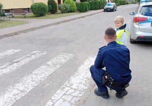 Droga- przejście dla pieszych. Po prawej przy drodze stoi dziecko w kamizelce odblaskowej. Przy nim kuca policjant.
