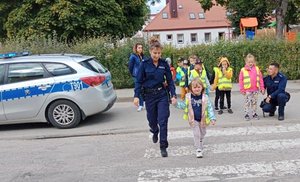 Droga- przejście dla pieszych. W tle po lewej stoi radiowóz.  Przez ulicę przechodzi policjantka trzymając dziecko za rękę. Po prawej w tle klęka policjant, który rozmawia z dzieckiem.