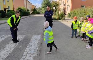 Droga - przejście dla pieszych. Po prawej na chodniku stopi grupka dzieci. Po lewej na drodze stoi policjantka w kamizelce odblaskowej. Przez drogę przechodzi dziecko.