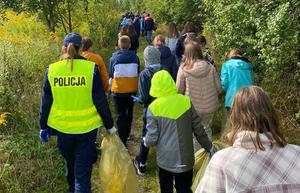 Droga w lesie. Ścieżką idzie grupa młodzieży odwrócona tyłem, a po lewej policjantka w kamizelce odblaskowej idąca razem z młodzieżą.