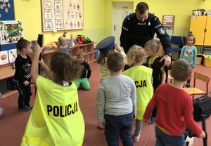 Sala w przedszkolu. Dzieci przymierzają policyjne kamizelki i czapki. Wśród nich widać policjanta.