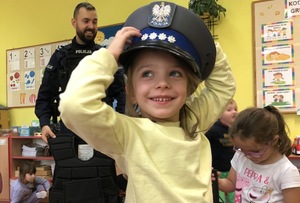 Na pierwszym planie uśmiechnięta dziewczynka w policyjnej czapce. W tle widać policjanta i przedszkolaki.