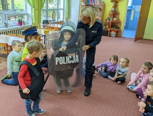 Sala w przedszkolu. Dzieci przymierzają policyjne kamizelki i czapki. Wśród nich policjantka,która pomaga dziecku trzymać policyjną tarczę.