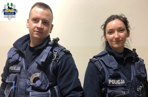 Na zdjęciu widać dwoje policjantów stojących obok siebie na tle jasnej ściany. Po lewej policjant, a po prawe policjantka.