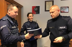 Po prawe policjant, który wręcza dokument dla policjanta po lewej. W tle uśmiechnięta policjantka.