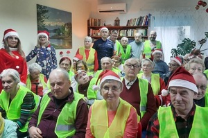 W grupie osób siedzących w sali w kamizelkach odblaskowych stoi policjant w mikołajowej czapce.